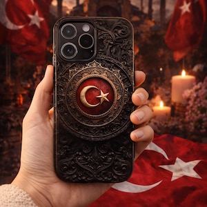 以下が含まれることがあります： トルコの国旗をあしらった、装飾的なデザインの黒い携帯電話ケース。ケースには詳細なエンボス加工の模様と赤と金のエンブレムがあります。背景にはトルコの国旗とぼやけた建築構造が含まれています。