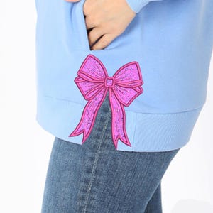 May include: Pale blue sweatshirt featuring a pink glitter bow appliqué. The bow is intricately designed with a textured finish. The sweatshirt is worn with blue denim jeans, offering a relaxed, casual style.
