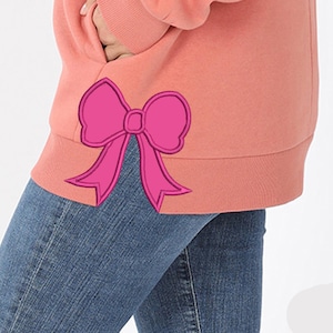 May include: A coral-colored sweatshirt with a large, bright pink bow applique on the lower left side. The bow is outlined in a darker pink, adding dimension. The sweatshirt is paired with blue denim jeans.
