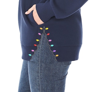 May include: Navy blue sweatshirt with a pocket and a decorative string of colorful lights embroidered along the hem. The sweatshirt is paired with blue jeans, creating a casual and festive look.