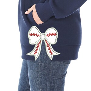 May include: Navy blue sweatshirt with a white baseball bow applique. The bow features red stitching details, resembling a baseball. The sweatshirt is paired with blue jeans.
