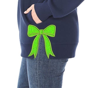 May include: Navy blue sweatshirt featuring a bright green bow design. The bow is embroidered on the lower left side of the sweatshirt. The person is wearing blue jeans. The sweatshirt has a pocket.
