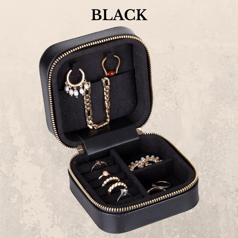 K&ouml;nnte beinhalten: Schwarzes Schmuck-Reiseetui mit goldenem Rei&szlig;verschluss und F&auml;chern. Das Etui ist ge&ouml;ffnet und zeigt verschiedene Schmuckst&uuml;cke, darunter Ohrringe, Ringe und eine Halskette. Das Wort "BLACK" ist oben im Bild aufgedruckt.