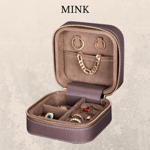 K&ouml;nnte beinhalten: Ge&ouml;ffnete, quadratische Schmuck-Reisetasche in Nerzfarbe mit goldenem Rei&szlig;verschluss und wei&szlig;en N&auml;hten. Der Innenraum ist mit einem weichen, hellbraunen Stoff ausgekleidet und enth&auml;lt verschiedene Ringe, Ohrringe und eine goldene Kette. Das Wort "MINK" ist oben aufgedruckt.