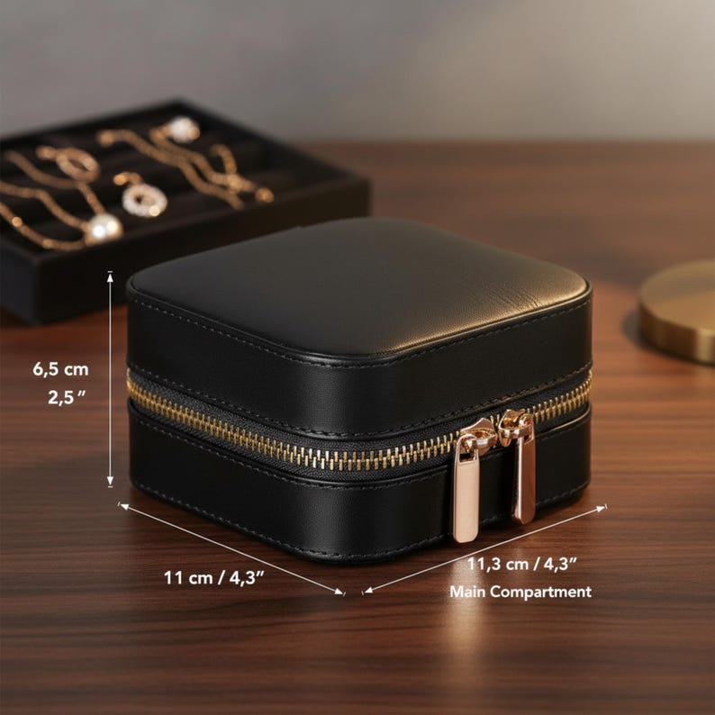 K&ouml;nnte beinhalten: Eine schwarze, quadratische Schmuckschatulle mit goldenem Rei&szlig;verschluss und Zug. Die Box ist 11 cm breit und 6,5 cm hoch. Im Hintergrund befindet sich ein schwarzes Schmucktablett mit goldenen Halsketten und Ohrringen.