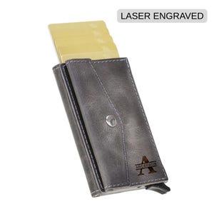May include: Gray leather card holder with a snap closure, holding several yellow cards. The text "LASER ENGRAVED" is at the top. The holder is personalized with the letter "A" and the name "ANDERSON".