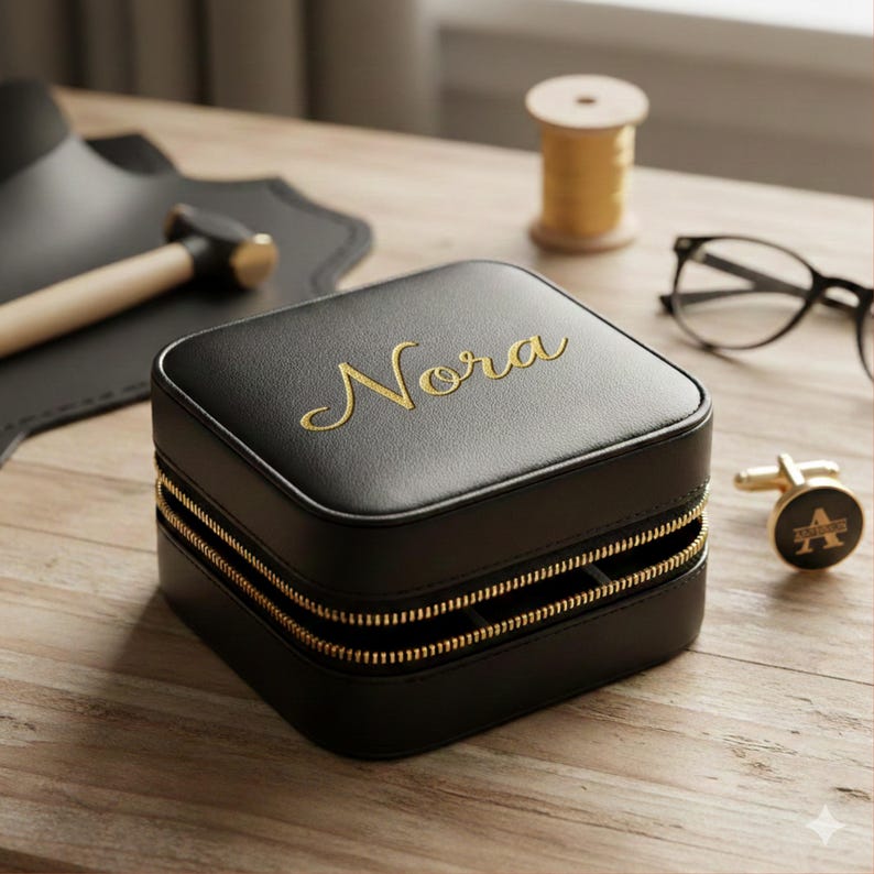K&ouml;nnte beinhalten: Eine schwarze, quadratische Schmuckschatulle mit goldenen Rei&szlig;verschl&uuml;ssen und dem Namen "Nora" in goldener Schrift. Ein goldfarbener Manschettenknopf mit dem Buchstaben "A" ist sichtbar. Die Box steht auf einer Holzoberfl&auml;che.