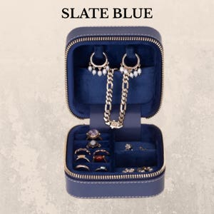 K&ouml;nnte beinhalten: Ge&ouml;ffnete, schieferblaue Schmuck-Reisetasche mit goldenem Rei&szlig;verschluss, die Ohrringe, Ringe und eine Halskette zeigt. Der Innenraum ist mit dunkelblauem Samt ausgekleidet. Der Text "SLATE BLUE" befindet sich oben im Bild.