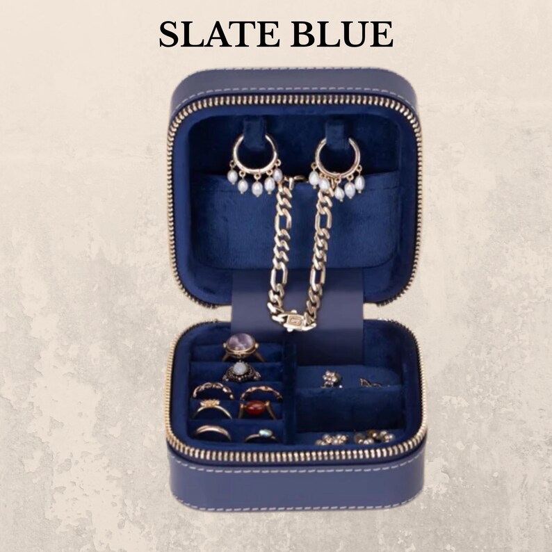 K&ouml;nnte beinhalten: Ge&ouml;ffnete, schieferblaue Schmuck-Reisetasche mit goldenem Rei&szlig;verschluss, die Ohrringe, Ringe und eine Halskette zeigt. Der Innenraum ist mit dunkelblauem Samt ausgekleidet. Der Text "SLATE BLUE" befindet sich oben im Bild.