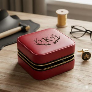K&ouml;nnte beinhalten: Eine rote, quadratische Schmuckschatulle mit goldenem Rei&szlig;verschluss und den Initialen "TKS" und Geweih auf der Oberseite. Ein goldener Manschettenknopf mit dem Buchstaben "A" befindet sich neben der Schatulle. Die Schatulle steht auf einer Holzoberfl&auml;che.