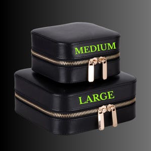 K&ouml;nnte beinhalten: Zwei schwarze quadratische Schmuckk&auml;stchen, eines mit der Aufschrift "MEDIUM" und das andere mit "LARGE". Beide Boxen haben goldfarbene Rei&szlig;verschl&uuml;sse und Ziehgriffe. Die Boxen sind vor dunklem Hintergrund gestapelt.