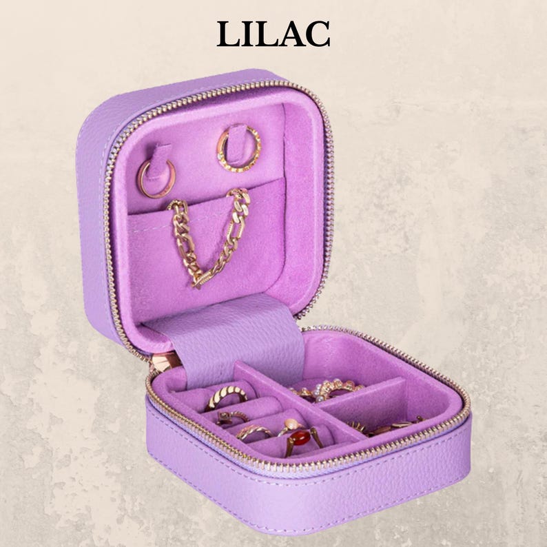 K&ouml;nnte beinhalten: Ein lila-farbenes, mit Rei&szlig;verschluss versehenes Schmuck-Reiseetui, das ge&ouml;ffnet ist, um verschiedene goldfarbene Ringe, Ohrringe und eine Kettenhalskette zu zeigen. Der Innenraum ist mit einem weichen, hellvioletten Stoff ausgekleidet. Das Wort "LILAC" ist oben aufgedruckt.