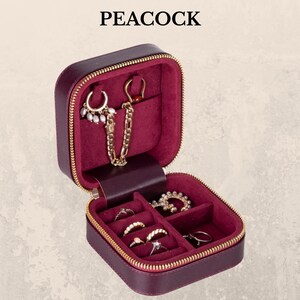 K&ouml;nnte beinhalten: Ein burgunderfarbenes Schmuck-Reiseetui mit goldenem Rei&szlig;verschluss und dem Wort "PEACOCK" oben. Der Innenraum ist mit burgunderfarbenem Samt ausgekleidet und bietet Platz f&uuml;r verschiedene Ringe und Ohrringe. Das Etui ist ge&ouml;ffnet und zeigt den Schmuck.