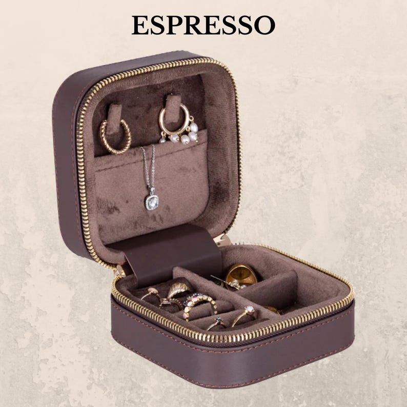 K&ouml;nnte beinhalten: Eine braune, mit Rei&szlig;verschluss versehene Schmuck-Reisetasche, ge&ouml;ffnet, um verschiedene Ringe, Ohrringe und eine Halskette zu pr&auml;sentieren. Der Innenraum ist mit einem weichen, braunen Stoff ausgekleidet. Das Wort "ESPRESSO" ist oben aufgedruckt. Die Tasche dient zur Aufbewahrung und zum Schutz von Schmuck.