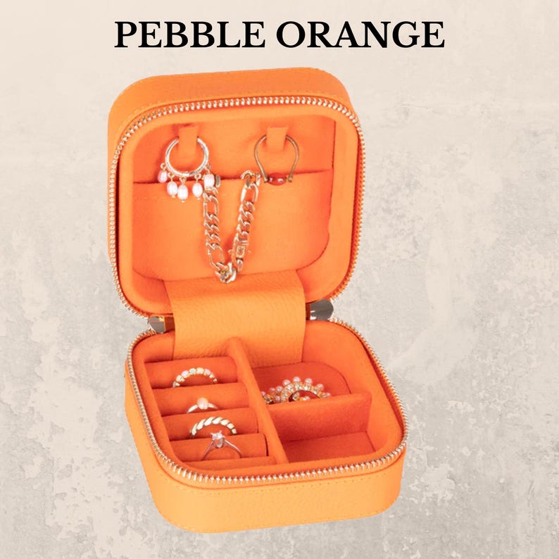 K&ouml;nnte beinhalten: Ge&ouml;ffnete orangefarbene Schmuck-Reisetasche mit Rei&szlig;verschluss, mit Ohrringen, Ringen und einer Kette. Auf der Tasche steht "PEBBLE ORANGE". Der Innenraum ist in F&auml;cher unterteilt.
