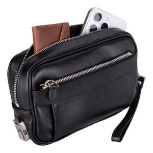 May include: Black leather clutch with a silver zipper and a wrist strap. The clutch is open, revealing a brown leather wallet and a white smartphone. The bag has a rectangular shape and a small pocket on the front.