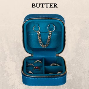K&ouml;nnte beinhalten: Ge&ouml;ffnete, t&uuml;rkisfarbene Schmuck-Reisetasche mit goldenem Rei&szlig;verschluss und dem Wort "BUTTER" oben. Der Innenraum verf&uuml;gt &uuml;ber ein t&uuml;rkisfarbenes Futter, eine goldene Kette und F&auml;cher f&uuml;r Ringe und Ohrringe. Die Tasche ist quadratisch und aus glattem Material.
