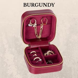 K&ouml;nnte beinhalten: Ge&ouml;ffnete, weinrote Schmuck-Reisetasche mit goldenem Rei&szlig;verschluss und Innenraum. Das Etui enth&auml;lt verschiedene Ringe, Ohrringe und eine goldene Kette. Das Wort "BURGUNDY" ist oben im Bild aufgedruckt.
