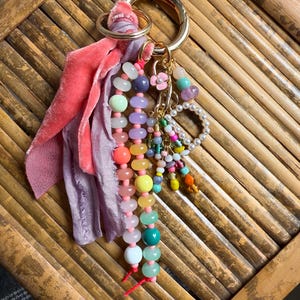 May include: A gold-toned metal keyring featuring a collection of multi-colored beads in various shapes and sizes, including translucent, opaque, and pearl-like finishes. Attached are strips of pink and lavender velvet fabric and a small flower charm.