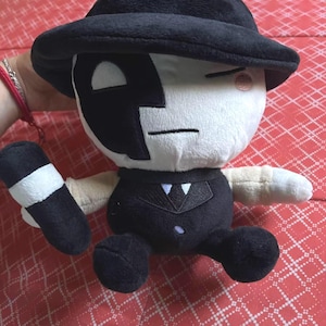 May include: A plush doll wearing a black hat, suit, and shoes. The doll has a white face with black markings and a black cylindrical object with a white band. It is on a red patterned surface.