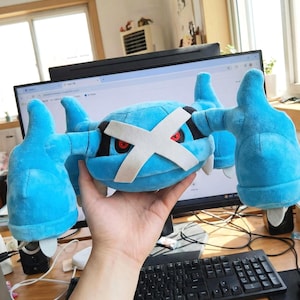 Op de afbeelding: Een blauw Metagross Pokémon knuffel, met een wit kruis en rode ogen. Het speelgoed wordt vastgehouden, met een computermonitor en toetsenbord op de achtergrond.