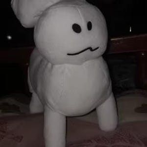 May include: A white plush bunny toy with black eyes and a curved mouth. The bunny has large, floppy ears and four short legs. The toy is made of soft material and is sitting on a patterned surface.