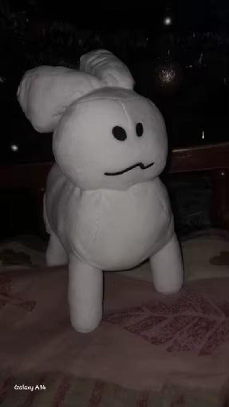 May include: A white plush bunny toy with black eyes and a curved mouth. The bunny has large, floppy ears and four short legs. The toy is made of soft material and is sitting on a patterned surface.