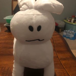 May include: A white plush bunny toy with large, floppy ears and a simple face. The bunny has two black oval eyes and a curved mouth. The toy is standing upright on a wooden surface.
