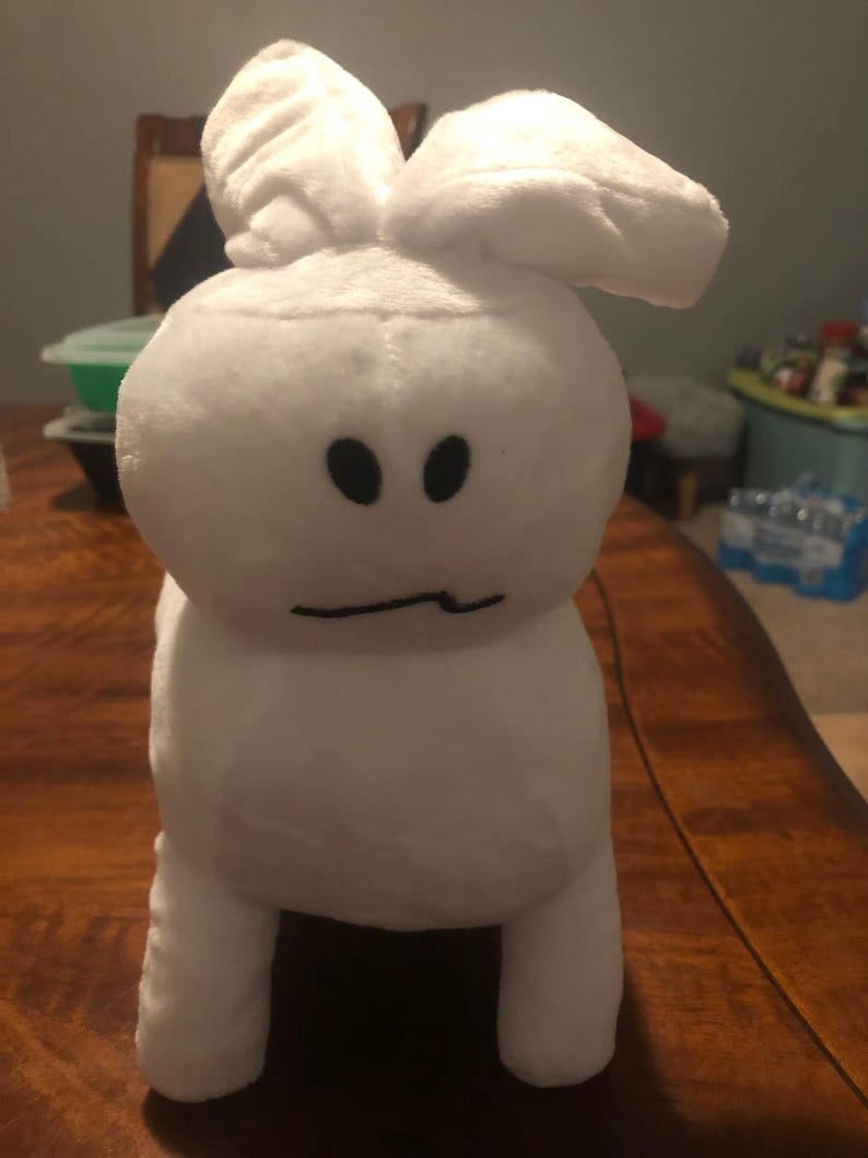 May include: A white plush bunny toy with large, floppy ears and a simple face. The bunny has two black oval eyes and a curved mouth. The toy is standing upright on a wooden surface.