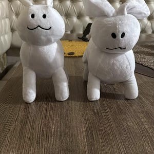 May include: Two white plush bunny toys with black embroidered facial features. The bunnies have upright ears and are positioned on a brown wooden surface. The background includes a cream-coloured sofa and decorative wall tiles.
