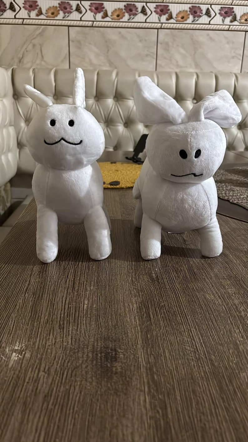 May include: Two white plush bunny toys with black embroidered facial features. The bunnies have upright ears and are positioned on a brown wooden surface. The background includes a cream-coloured sofa and decorative wall tiles.
