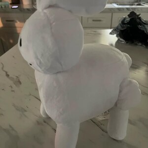 May include: A white plush bunny toy with a rounded head, long ears, and four stubby legs. The bunny has small black eyes and is made of a soft, fluffy material. The toy is standing on a light-coloured surface.