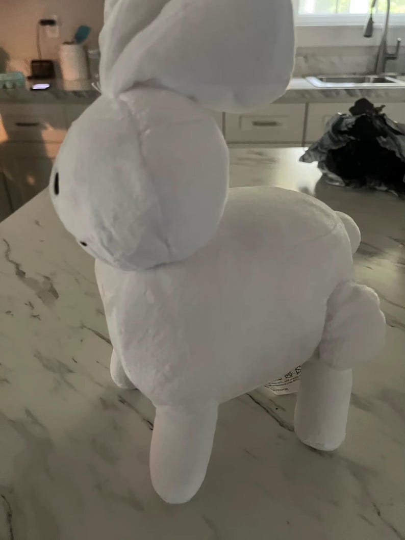 May include: A white plush bunny toy with a rounded head, long ears, and four stubby legs. The bunny has small black eyes and is made of a soft, fluffy material. The toy is standing on a light-coloured surface.