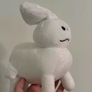 May include: A white plush bunny toy with a minimalist design. The bunny has a rounded body, small legs, and long ears. It features a simple black line for a mouth and two black dots for eyes. The toy is held in a hand.