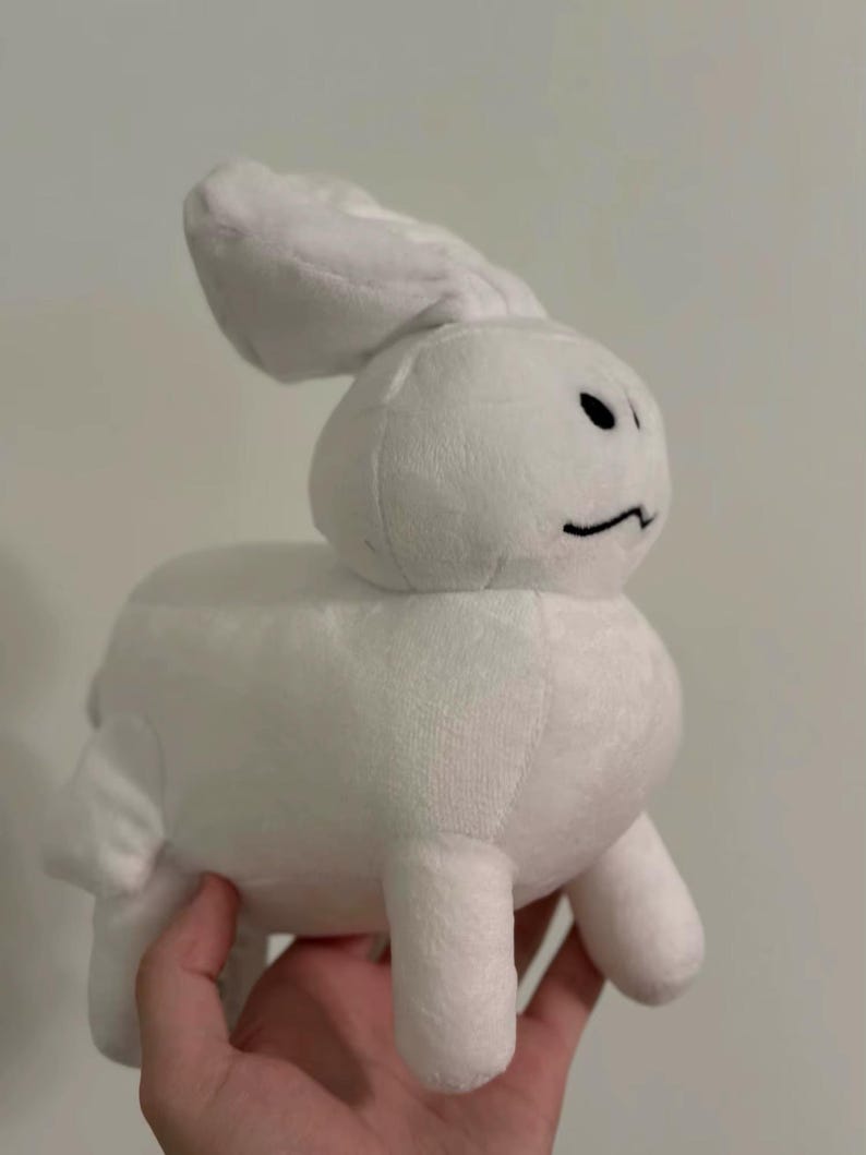 May include: A white plush bunny toy with a minimalist design. The bunny has a rounded body, small legs, and long ears. It features a simple black line for a mouth and two black dots for eyes. The toy is held in a hand.
