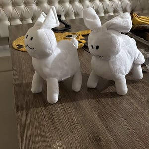 May include: Two white plush bunny rabbit toys with black stitched features. The rabbits have long ears and four legs. They are positioned on a brown wooden surface.