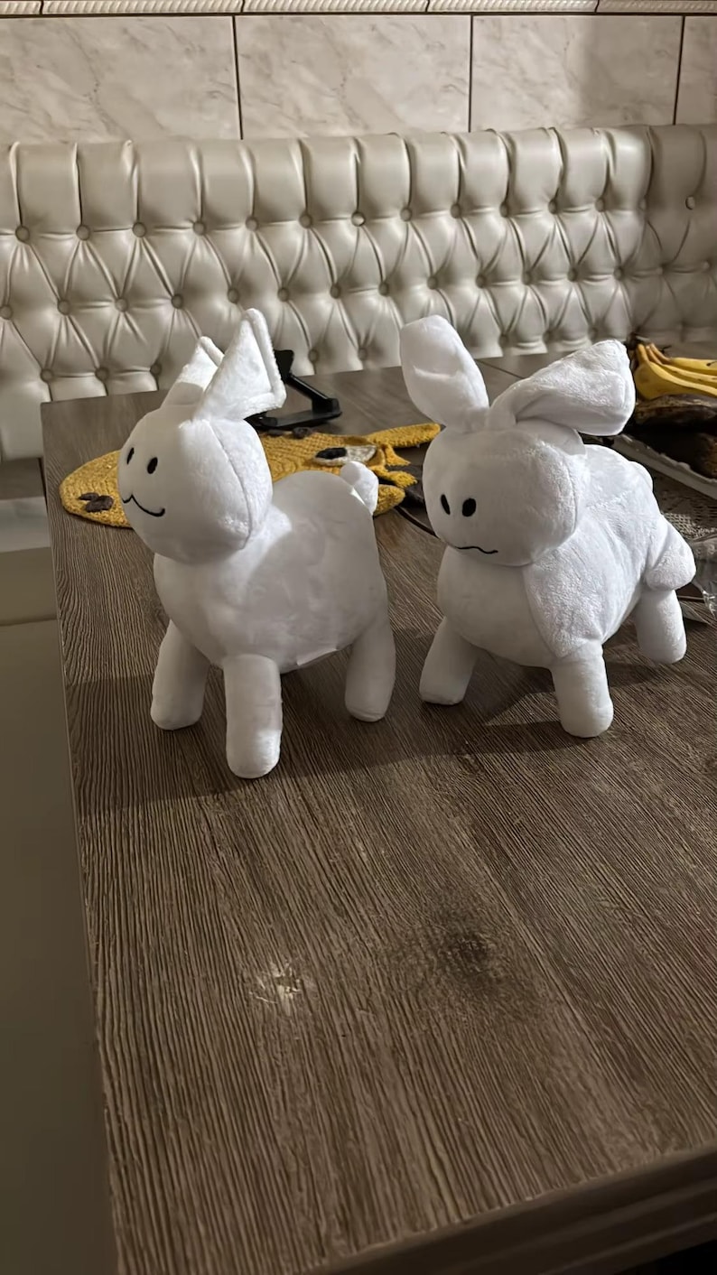 May include: Two white plush bunny rabbit toys with black stitched features. The rabbits have long ears and four legs. They are positioned on a brown wooden surface.