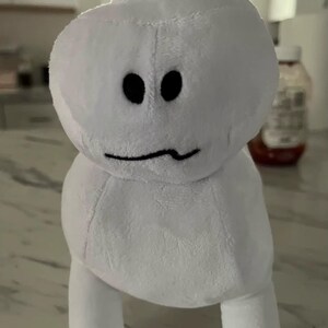 May include: A white plush toy with a rounded body, two small legs, and a head featuring two black eyes and a curved mouth. The toy has a simple, cartoonish design and a soft, fuzzy texture.
