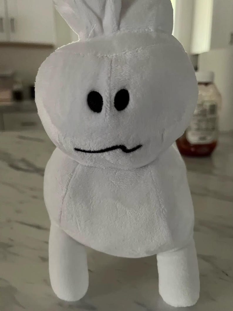 May include: A white plush toy with a rounded body, two small legs, and a head featuring two black eyes and a curved mouth. The toy has a simple, cartoonish design and a soft, fuzzy texture.