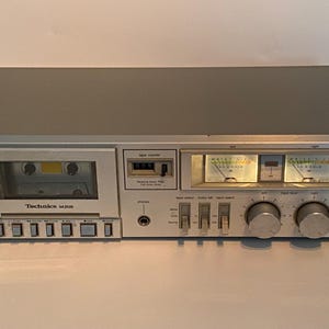 May include: Silver Technics M205 cassette deck with a tape counter, VU meters, and various control knobs and buttons. The cassette player has a tape inserted and the Technics logo is visible.