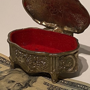 May include: A silver trinket box with an ornate floral design and three legs. The box is open and has a red velvet interior.