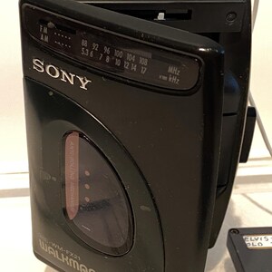 May include: Black Sony Walkman FM/AM radio, model WM-FX21, with the door open. The radio has a tuning dial and a headphone jack. The text "SONY" is visible on the front. The radio is connected to a white cable.