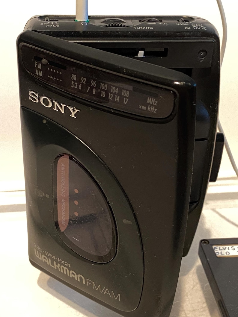 May include: Black Sony Walkman FM/AM radio, model WM-FX21, with the door open. The radio has a tuning dial and a headphone jack. The text "SONY" is visible on the front. The radio is connected to a white cable.
