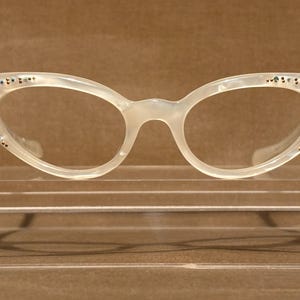 May include: Cat-eye glasses with a translucent off-white pearlescent frame. The outer corners of the frames are decorated with small, dark, circular embellishments arranged in a starburst pattern on each side of the eyewear.