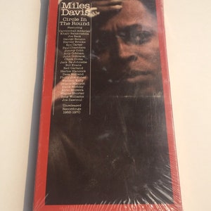 May include: A red and black CD case with a photo of Miles Davis on the front. The case is titled "Miles Davis Circle In The Round" and features a list of musicians including Cannonball Adderley, Khalil Balakrishna, Joe Beck, George Benson, Harvey Brooks, and more. The case also includes the text "Digitally Remastered Directly From The Original Analog Tapes" and "Columbia Jazz Contemporary Masters".
