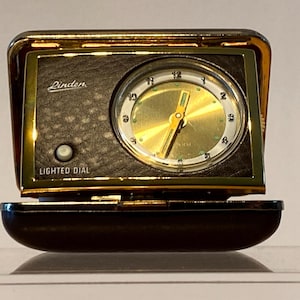 May include: A gold-colored Linden brand travel alarm clock with a black face and a lighted dial feature. The clock has a gold-colored metal case and a black leatherette base.
