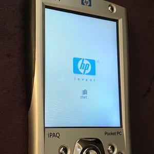 May include: Silver HP iPAQ Pocket PC with a bright screen displaying the HP logo and the word "invent". The device has a central navigation button and several smaller buttons below the screen. The device is a handheld computer.