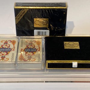 May include: Two decks of Congress Playing Cards in a clear plastic case. The cards are plastic coated and feature a joker design. The case has a black velvet lined drawer with a gold trim.