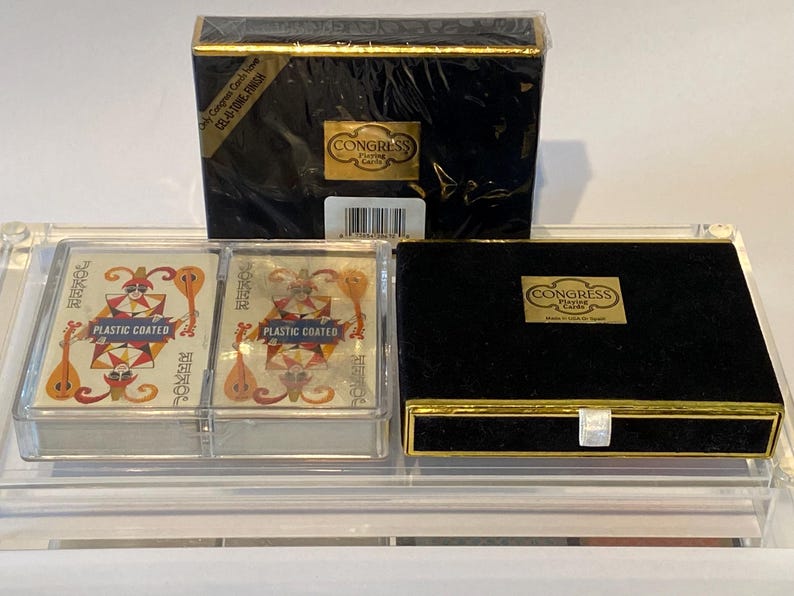 May include: Two decks of Congress Playing Cards in a clear plastic case. The cards are plastic coated and feature a joker design. The case has a black velvet lined drawer with a gold trim.
