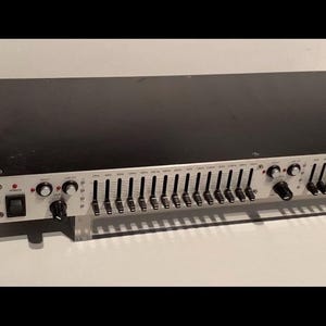 May include: A RadioShack audio equalizer with a black top and a silver face. It features numerous sliders, knobs, and buttons for adjusting sound frequencies. The RadioShack logo is visible on the left side.